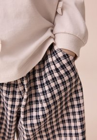 Sweat-shirt beige à encolure arrondie avec un détail de nœud sur la manche, associé à un pantalon imprimé à carreaux en tons noir et beige.