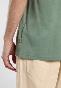 Grøn bomulds T-shirt med blød tekstur. Sidevisning viser syninger og et lille logo mærke. Sættes sammen med lyse beige shorts med lommer.