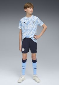 Zils un balts svītrainā īssvītra futbola krekls, melni šorti, zili un tumši zili zeķes, baltas futbola kurpes, ar Puma logo un Mančestra City emblēmu.