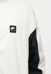 Λευκό και μαύρο μακρυμάνικο μπουφάν Nike Air με το λογότυπο της Nike στο στήθος και μαύρο πάνελ στο μανίκι.