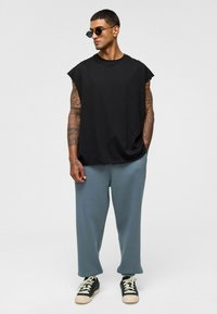 Zwarte mouwloze katoenen shirt gecombineerd met losse, grijze sweatpants en zwarte sneakers. Extra details zijn zonnebrillen en zichtbare tatoeages.