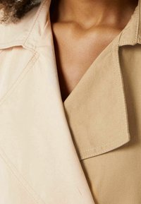 Beżowy płaszcz z wyłogowym kołnierzem, charakteryzujący się gładką fakturą i widocznymi detalami stawów. Materiał wydaje się lekki i miękki.