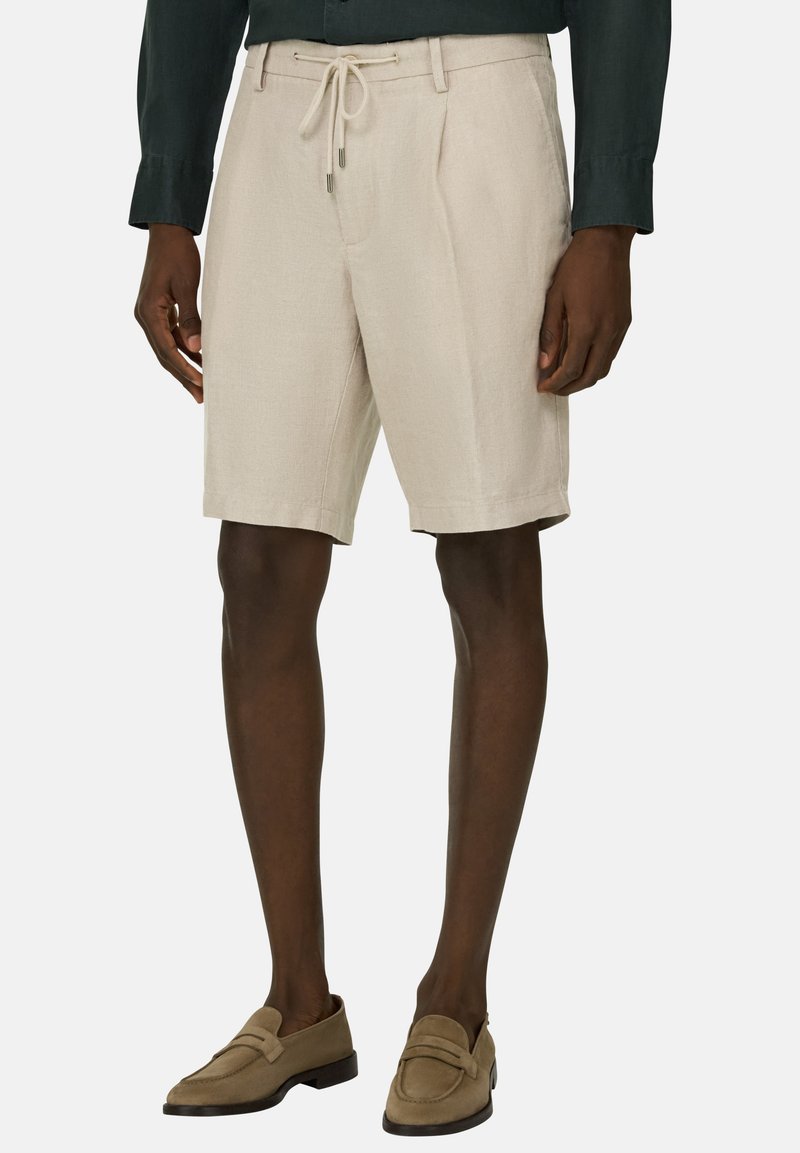 Homme portant un short beige mi-genou avec une taille à cordon, une chemise à manches longues foncée, et des mocassins beiges, debout devant un fond uni.