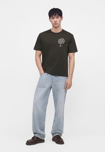 Νέος άνδρας με μαύρο t-shirt με λευκό σπειροειδές σχέδιο, ανοιχτό μπλε τζιν με φαρδιά μπατζάκια και σκούρα καφέ παπούτσια, στέκεται με το ένα χέρι στην τσέπη.