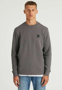 Felpa grigia a coste con collo rotondo, maniche lunghe e un piccolo emblema nero; abbinata a jeans azzurri chiari. Texture liscia.