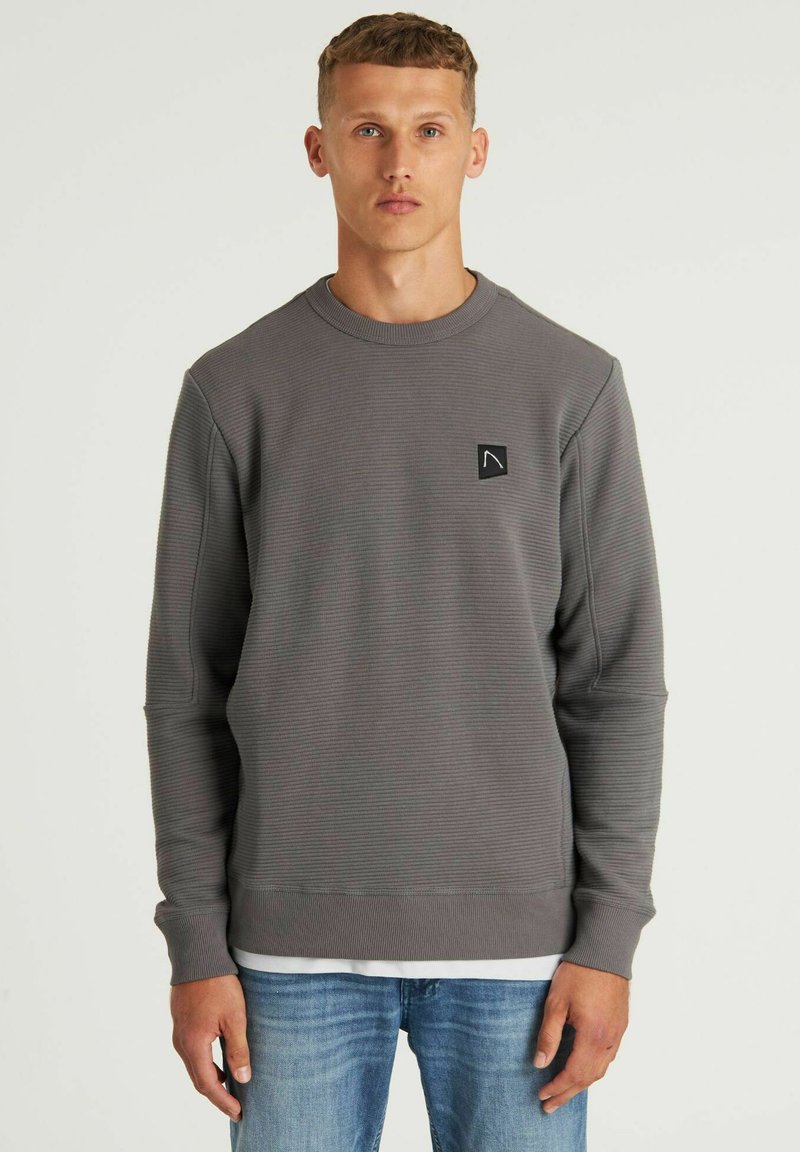 Felpa grigia a coste con collo rotondo, maniche lunghe e un piccolo emblema nero; abbinata a jeans azzurri chiari. Texture liscia.