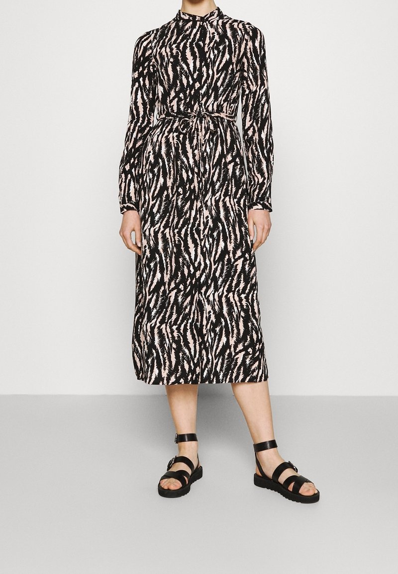 Femme portant une robe midi à manches longues, noire et rose pâle avec un motif abstrait, ceinturée à la taille, et des sandales noires à bride à la cheville.