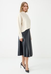 Maglione a maglia crema con polsini a coste, abbinato a una gonna plissettata in ecopelle nera. Modella che indossa tacchi a punta neri, in piedi contro uno sfondo bianco.