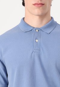 Lys blå polo skjorte laget av teksturert stoff, med klassisk krage og to knapper ved halsen, vist iført av en person.