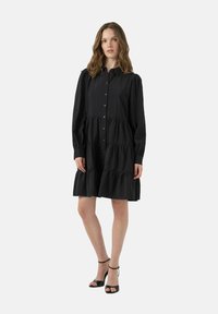 Abito nero con bottoni, maniche a sbuffo lunghe, design a strati con volant e tessuto leggero. Abbinato a sandali neri con cinturino.