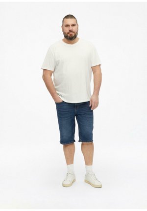 Man met baard die een wit t-shirt, blauwe denim shorts, witte sokken en witte sneakers draagt, staat met één hand in de zak op een witte achtergrond.