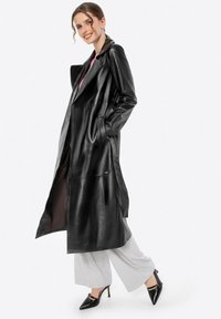 Zwarte leren trenchcoat met een brede kraag en zijzakken. Het heeft een lange, vloeiende uitstraling en wordt open gedragen over grijze broeken en hakken.