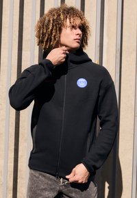 Sort zip-up hoodie med høj krave, der har et blåt cirkulært logo på brystet, tekstureret stof og sidelommer.