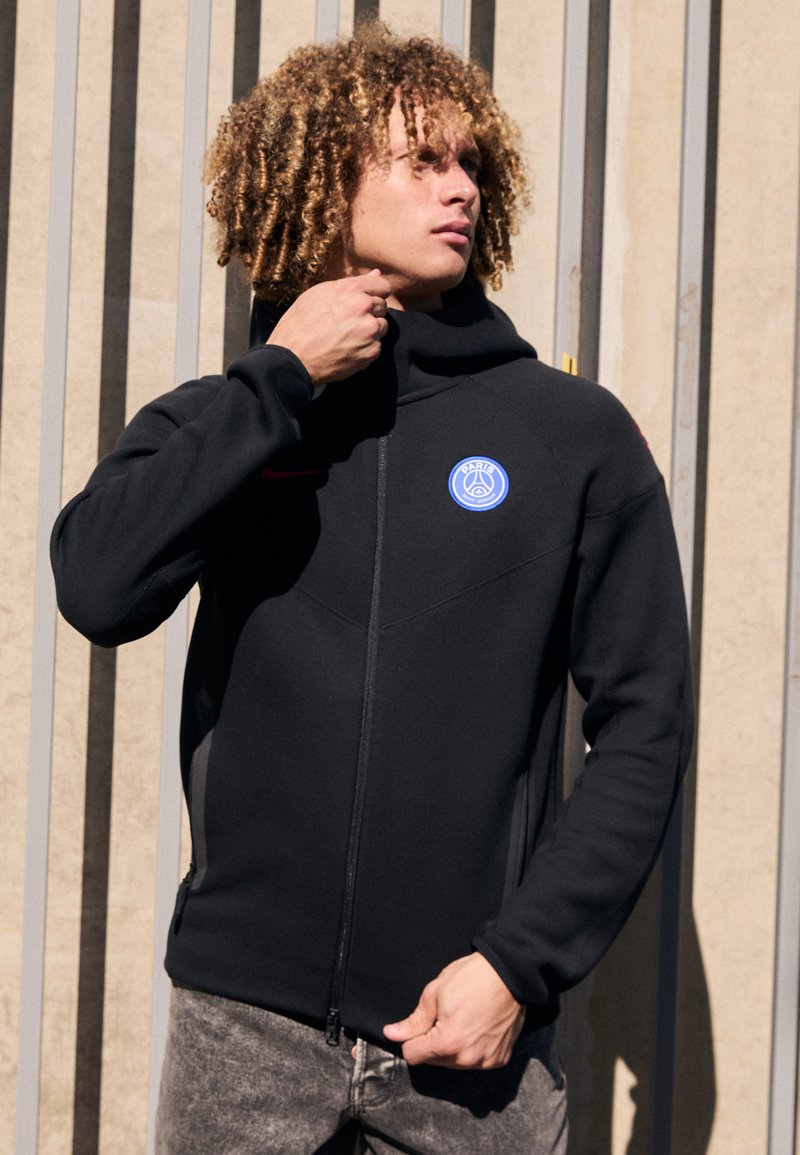 Sort zip-up hoodie med høj krave, der har et blåt cirkulært logo på brystet, tekstureret stof og sidelommer.