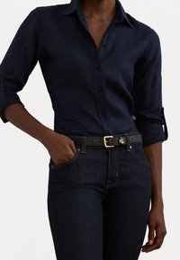 Marineblå skjorta med knappar i linne, med uppvikta ärmar, kombinerad med mörka denimjeans och ett svart bälte med ett guldspänne.