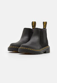 Bottes de cheville en cuir noir avec panneaux élastiques sur les côtés, coutures jaunes et languettes de tirage avec logo. Design épuré avec une semelle rugueuse et texturée.