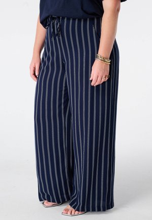 Persoon die marineblauwe wijde broek draagt met witte verticale strepen, een zwart topje, gouden armbanden en sandalen met open tenen.