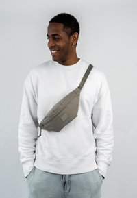 Borsa a tracolla grigia realizzata in tessuto resistente, dotata di chiusura con zip, tracolla diagonale e un'etichetta logo rettangolare, indossata sopra un sudore bianco.
