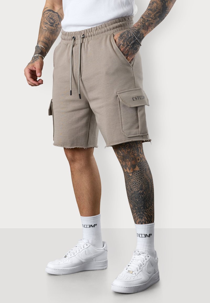 Shorts cargo beiges avec taille élastique, deux poches latérales et une poche avec l'étiquette "ESTEEM". Portés avec des baskets blanches et des chaussettes.
