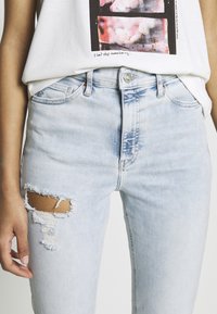 Jeans de denim azul claro con un diseño desgastado, que presentan un gran desgarro en el muslo izquierdo y un estilo clásico de cinco bolsillos.