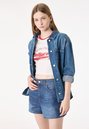 Jonge vrouw draagt een oversized spijkerjack, een witte cropped top met rode letters en hooggetailleerde spijkershorts, poserend tegen een witte achtergrond.