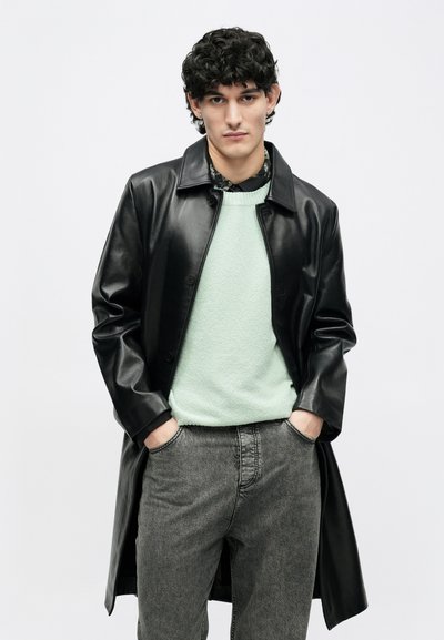 Abrigo negro de cuero con botones delanteros, llevado sobre un suéter verde claro. El modelo lleva jeans grises, mostrando un conjunto casual y en capas.