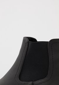 Bottine noire avec une texture lisse, panneau latéral élastique et un détail de couture. Design simple avec un bout rond.