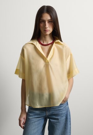 Junge Frau mit langen dunklen Haaren trägt eine durchsichtige, hellgelbe kurzärmelige Bluse, eine rote Perlenkette und blaue Jeans und posiert mit einer Hand in der Tasche.