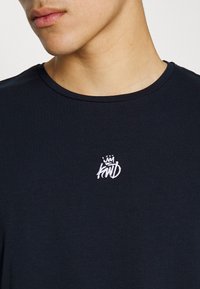 Black cotton t-shirt with a round neckline and short sleeves. Features white embroidered logo on the left chest area. Smooth texture.