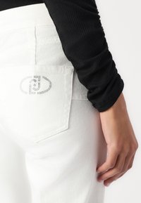 Biele džínsy z denimovej tkaniny s vreckom na zadnej strane a strieborným logom. Osoba má na sebe čierny top s dlhým rukávom, na fotke je zachytená časť ramena a ruky.