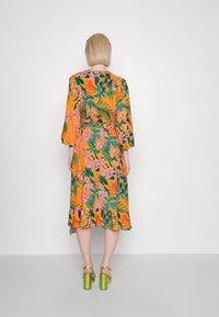 Kwiatowa sukienka w pomarańczowej podstawie, zdobiona różowymi i zielonymi wzorami liści. Długie rękawy, ściśnięta talia i falbana na dolnej krawędzi. Noszona z zielonymi szpilkami.