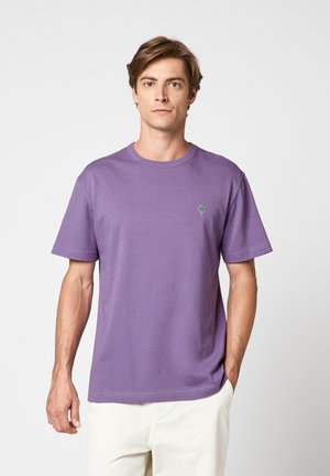 Homme portant un T-shirt violet uni à col rond avec un petit logo d'arbre vert sur la poitrine gauche et un pantalon blanc, debout avec une main dans la poche.