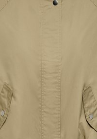 Veste beige devant avec fermeture à boutons-pression et deux poches à rabat sécurisées par des boutons-pression métalliques.
