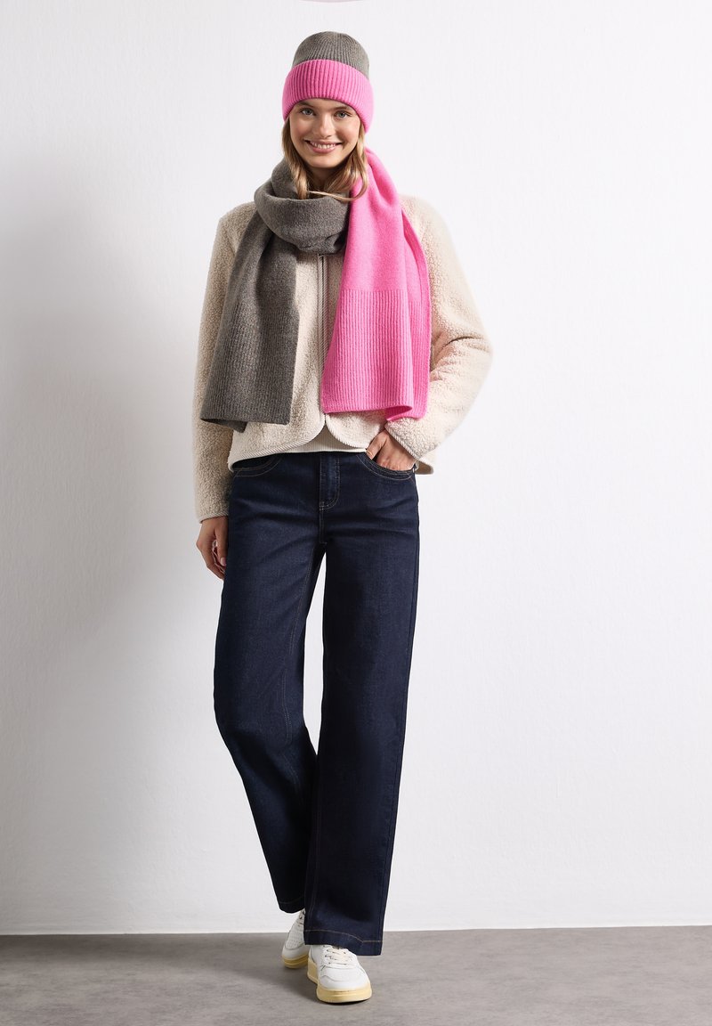 Jeune femme souriante, portant une veste polaire beige, un jean foncé, des baskets blanches, un bonnet bicolore gris et rose, et une écharpe assortie.