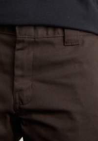 Pantalon en coton marron à texture lisse, présentant une coupe classique, des passants pour ceinture et des détails de coutures discrètes sur les poches.