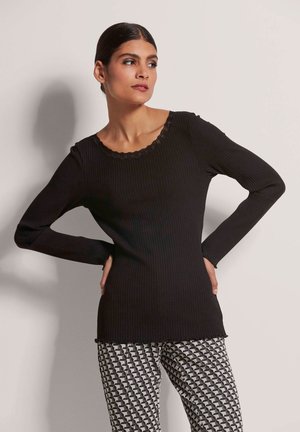 Schwarzes, geripptes Langarmshirt mit gewelltem Spitzenkragen. Kombiniert mit gemusterten schwarz-weißen geometrischen Hosen.