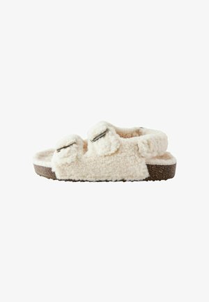 Chaussons crémeux et duveteux de couleur crème, avec des sangles ajustables doubles et une semelle en liège texturé. L'extérieur doux ressemble à du tissu en peau de mouton.