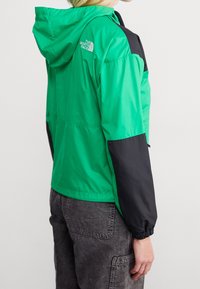 Zeleno-černá větrovka s kapucí, elastickými manžetami a lehkým designem, s logem na rameni.