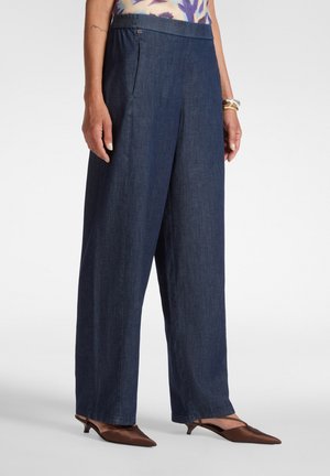 Frau trägt lockere dunkelblaue Jeanshosen mit Seitentaschen, ein gemustertes Oberteil und braune spitze Kitten Heels, steht vor einem schlichten Hintergrund.