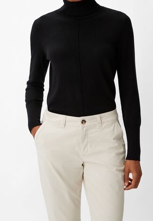 Personne portant un pull à col roulé noir rentré dans un pantalon beige taille haute, les mains partiellement dans les poches.
