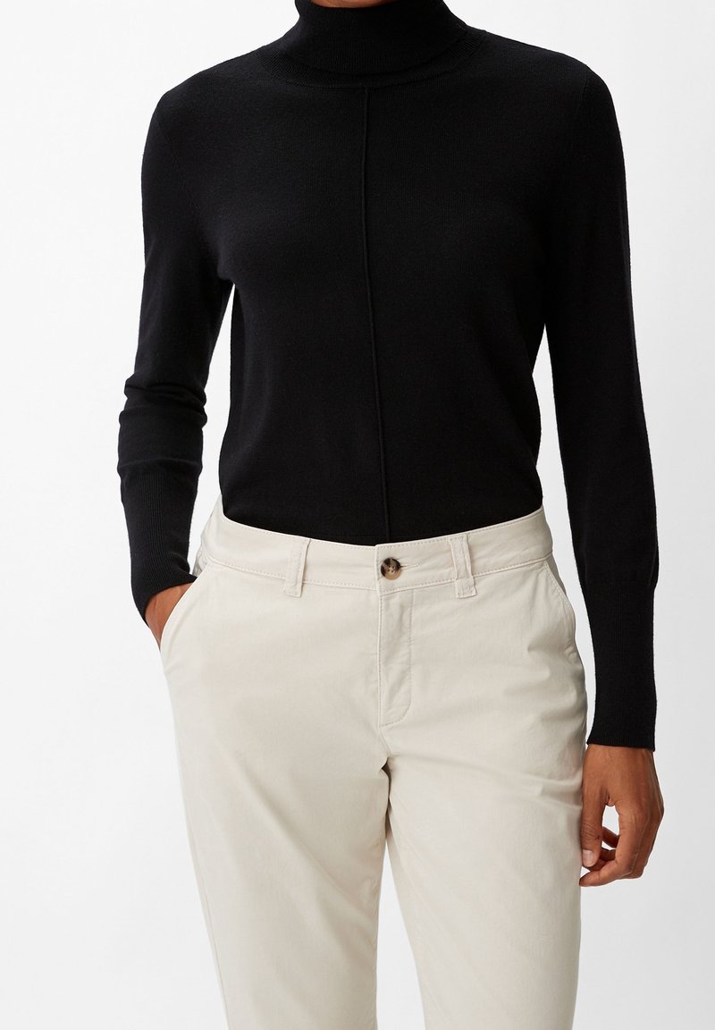 Personne portant un pull à col roulé noir rentré dans un pantalon beige taille haute, les mains partiellement dans les poches.