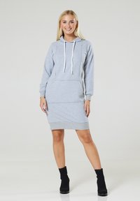 Graues Kapuzenkleid aus weichem Stoff, mit einer Vordertasche, geripptem Saum und verstellbaren Kordeln. Kombiniert mit schwarzen Knöchel-Stiefeln.