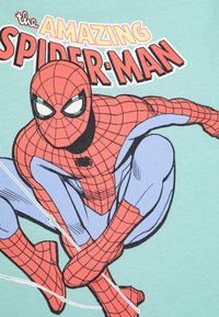 Žalia marškinėliai su komiksų herojumi Žmogumi voru, vilkinčiu raudoną ir mėlyną kostiumą, viršuje esantys žodžiai "NUOSTABUSIS ŽMOGUS VORAS" drąsiais rašmenimis.