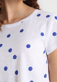 Camiseta de algodón blanca con lunares azules, con un cuello redondo y mangas cortas. Textura suave y diseño casual.