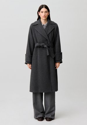 Donna con capelli scuri e lisci indossa un lungo cappotto grigio scuro con cintura sopra pantaloni larghi grigi e scarpe scure, in piedi su sfondo grigio chiaro.