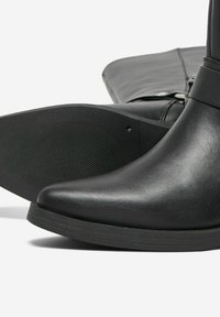 Bottine noire en cuir lisse avec un bout pointu. Elle dispose d'une semelle noire texturée et d'un accent de boucle réglable.
