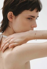 Joven con el cabello corto y oscuro, descansando la mano en el hombro, lleva una delicada pulsera de oro y dos anillos finos de oro con pequeñas piedras de colores.