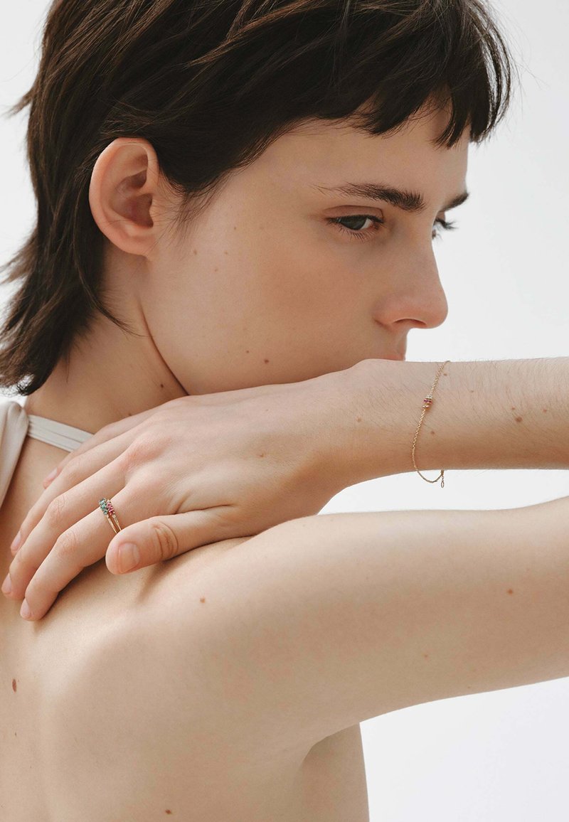 Joven con el cabello corto y oscuro, descansando la mano en el hombro, lleva una delicada pulsera de oro y dos anillos finos de oro con pequeñas piedras de colores.