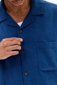 Camicia di lino blu con texture, colletto, bottoni in legno e una tasca sul petto, mostrata con una mano che sistema il bottone.