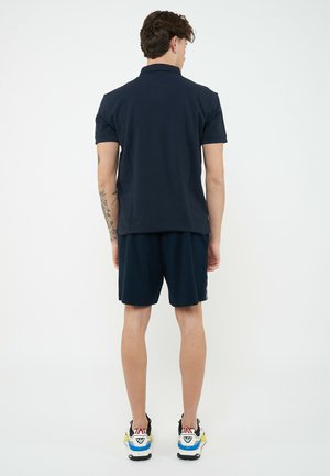 Hombre con camisa polo azul marino y pantalones cortos, de pie de espaldas con tatuaje visible en el brazo y zapatillas coloridas sobre un fondo blanco.